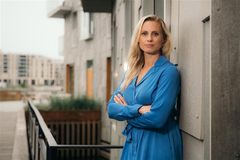 Silje Sandmæl iført blå kjole, lener seg mot husvegg, ser alvorlig ut