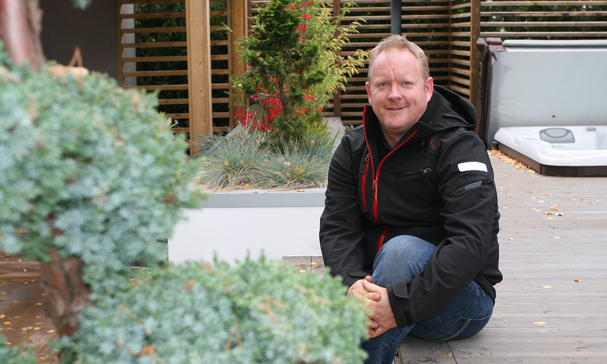 Darren Saines sitter på et terrassegulv iført sort allværsjakke. Smiler.