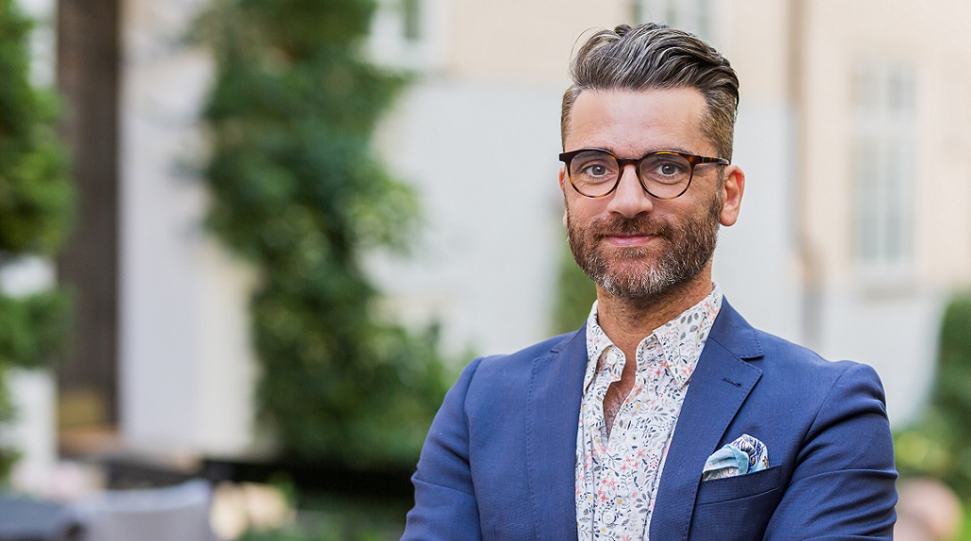 Megleren ser i kameraet og smiler. Han har på seg blå dressjakke med skjorte under