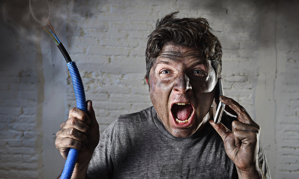 young man holding electrical cable smoking after domestic accident with dirty burnt funny face expression calling desparate with mobile phone asking for help in electricity DIY wrong repairs concept 